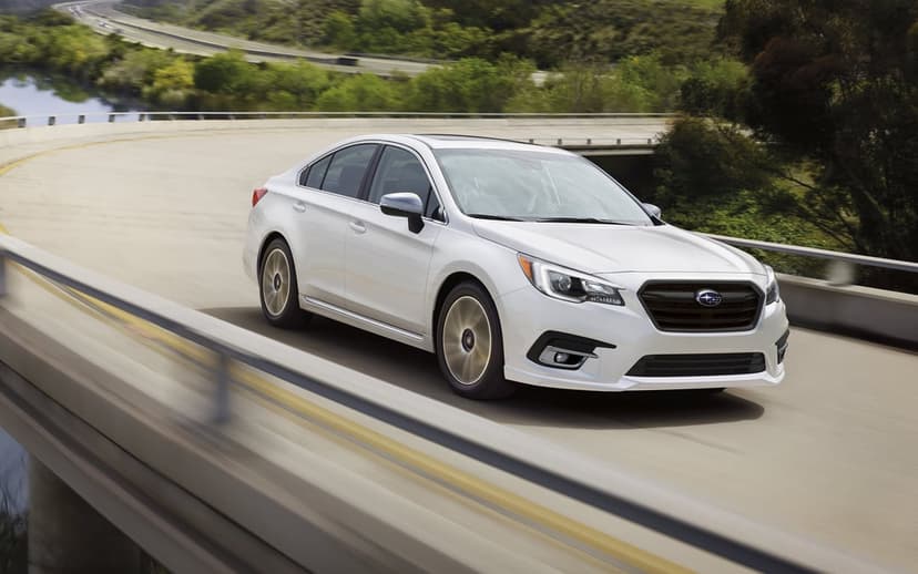 White Subaru sedan driving on a curved concrete bridge with motion blur against green hills and water.