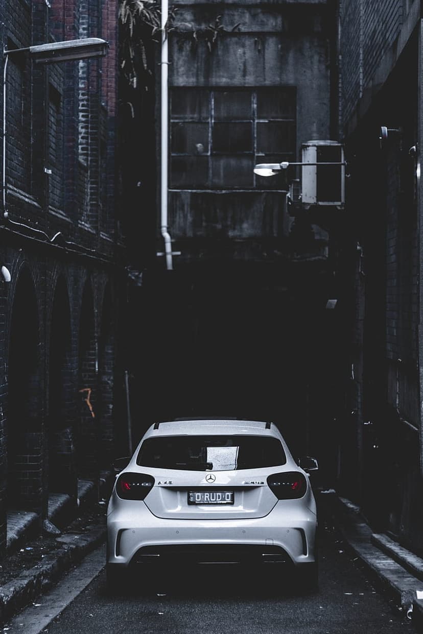 White Mercedes-Benz hatchback with lit taillights parked between dark brick walls in a narrow city alleyway.