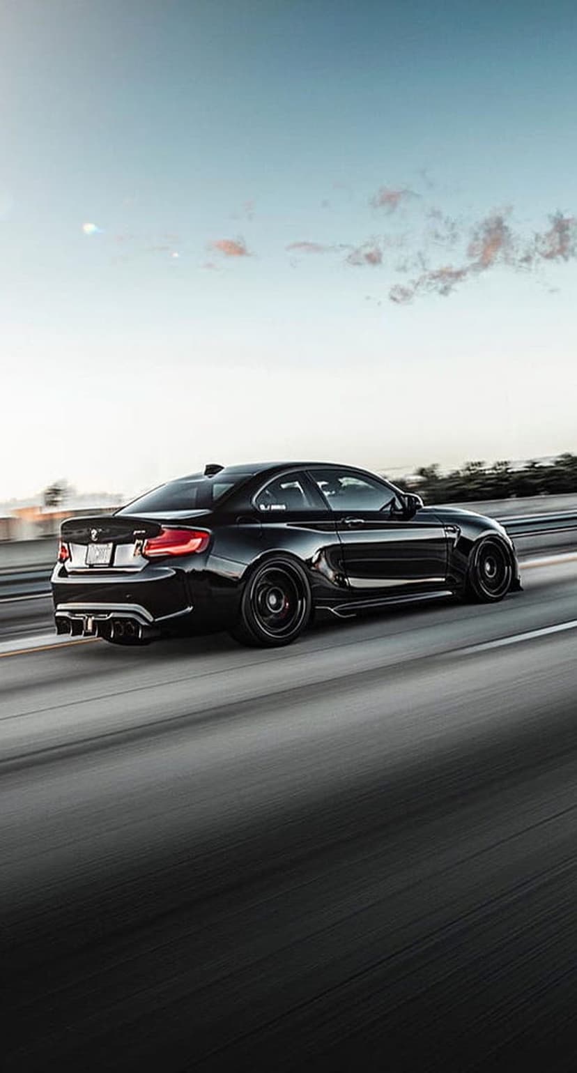 Black BMW M2 coupe drives on a highway at sunset with motion blur, wide body kit, and a low angle perspective.