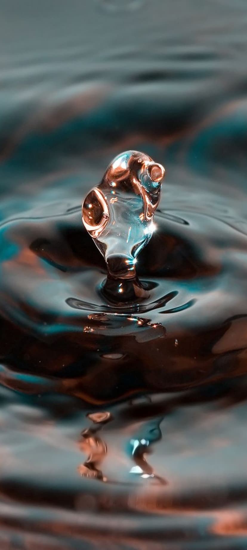 Golden water droplet frozen in a sculpture shape above dark rippling water with teal and brown reflections.