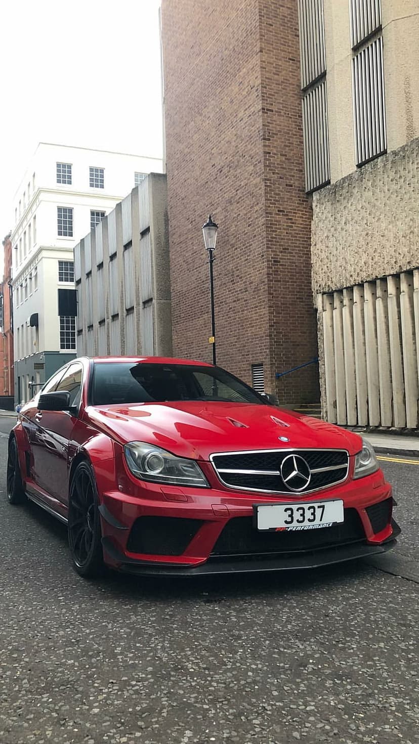 Red Mercedes AMG C63 with widebody fenders and black wheels parked on an asphalt city street near concrete buildings.