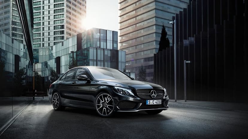 Black Mercedes-Benz AMG sedan parked in a modern city with glass skyscrapers and bright sun flare.