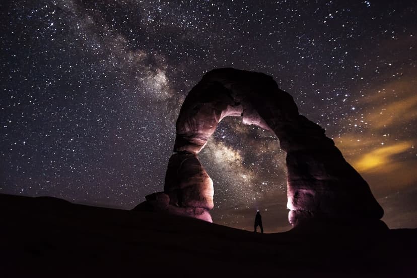 Lone person silhouetted beneath a sandstone arch under a starry Milky Way galaxy in the Utah desert at night.