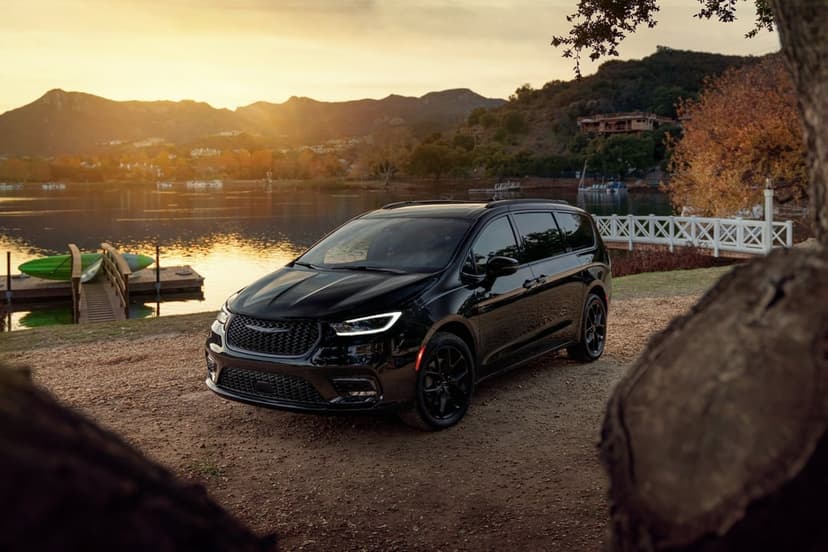 Black minivan on a dirt path by a lake at sunset with mountains and a wooden pier in the background.