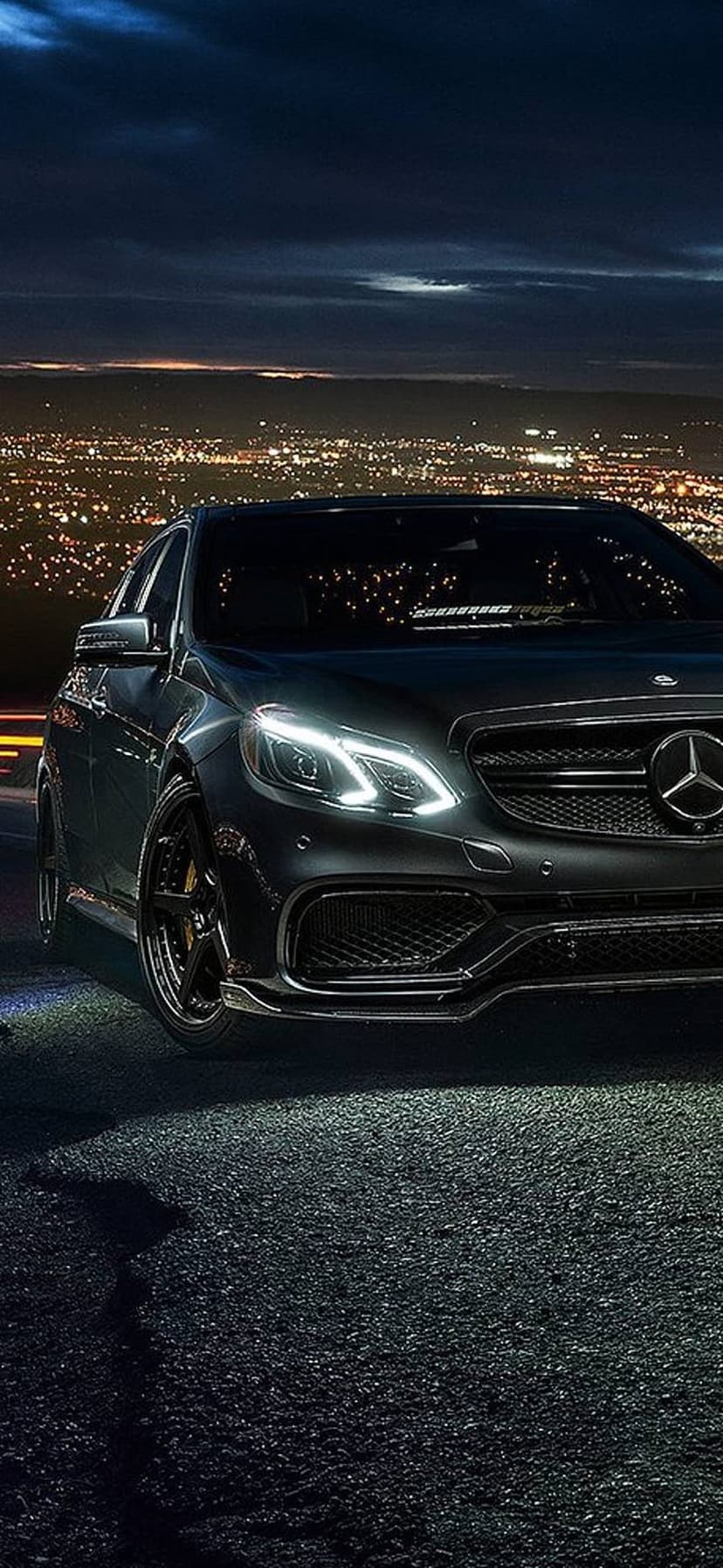 Black Mercedes-Benz E-Class sedan parked on asphalt with glowing headlights and a twinkling city backdrop.