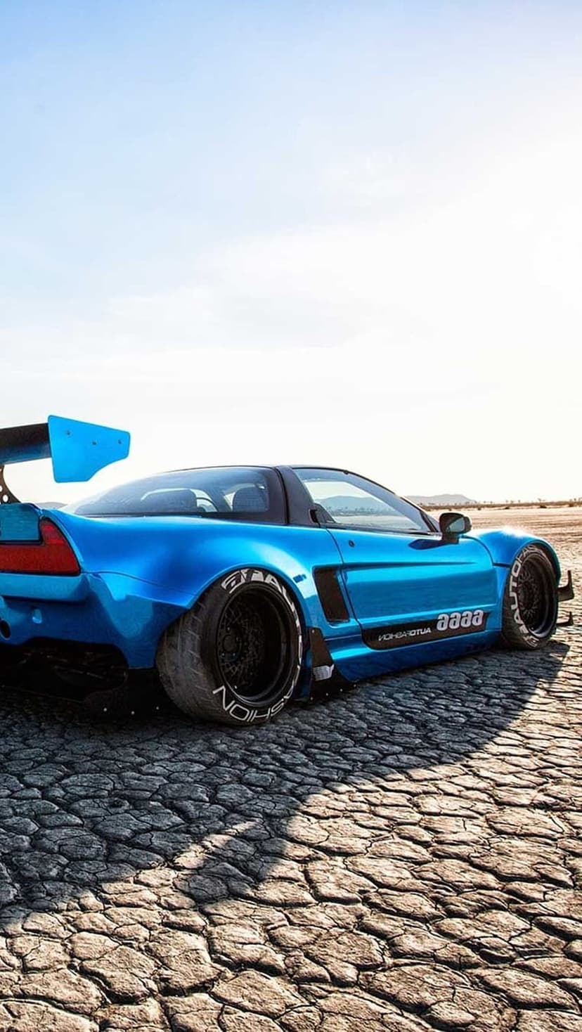 Blue Honda NSX with a widebody kit and rear wing parked on a dry, cracked desert floor under a clear sky.