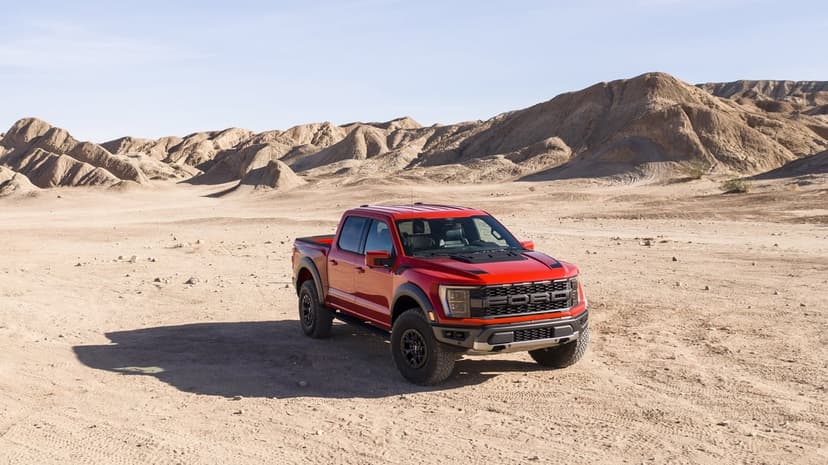 Red Ford F-150 Raptor truck parked on sandy ground with desert hills and a blue sky in the background.