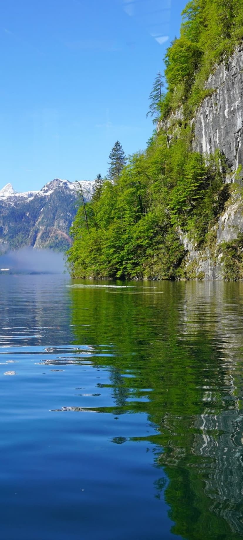 Emerald green cliffs and snow-capped peaks reflect on the calm, glassy surface of a crystal blue alpine lake.