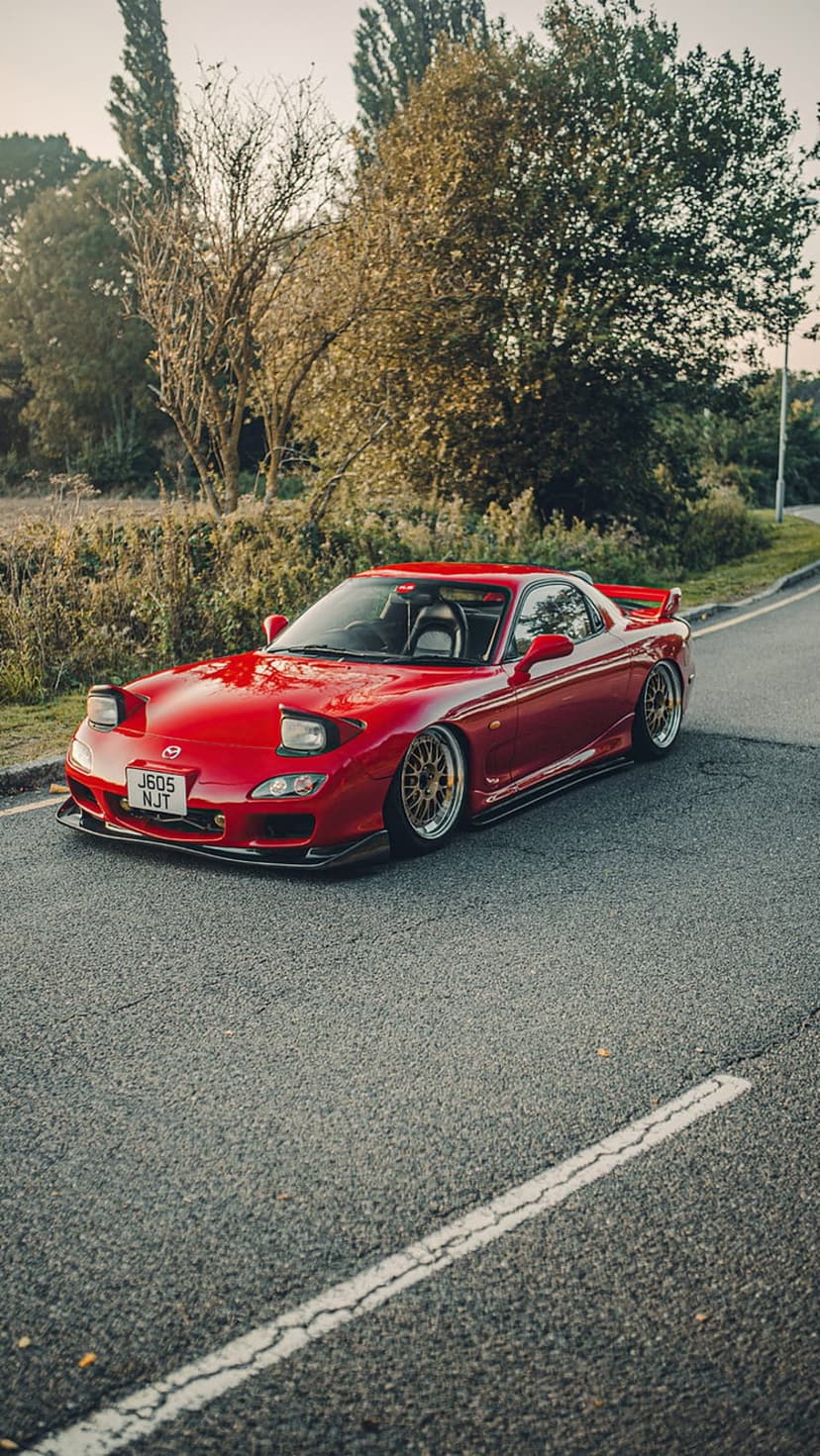 Red Mazda RX-7 FD parked on a leaf-strewn country road surrounded by orange autumn trees and golden sunlight.