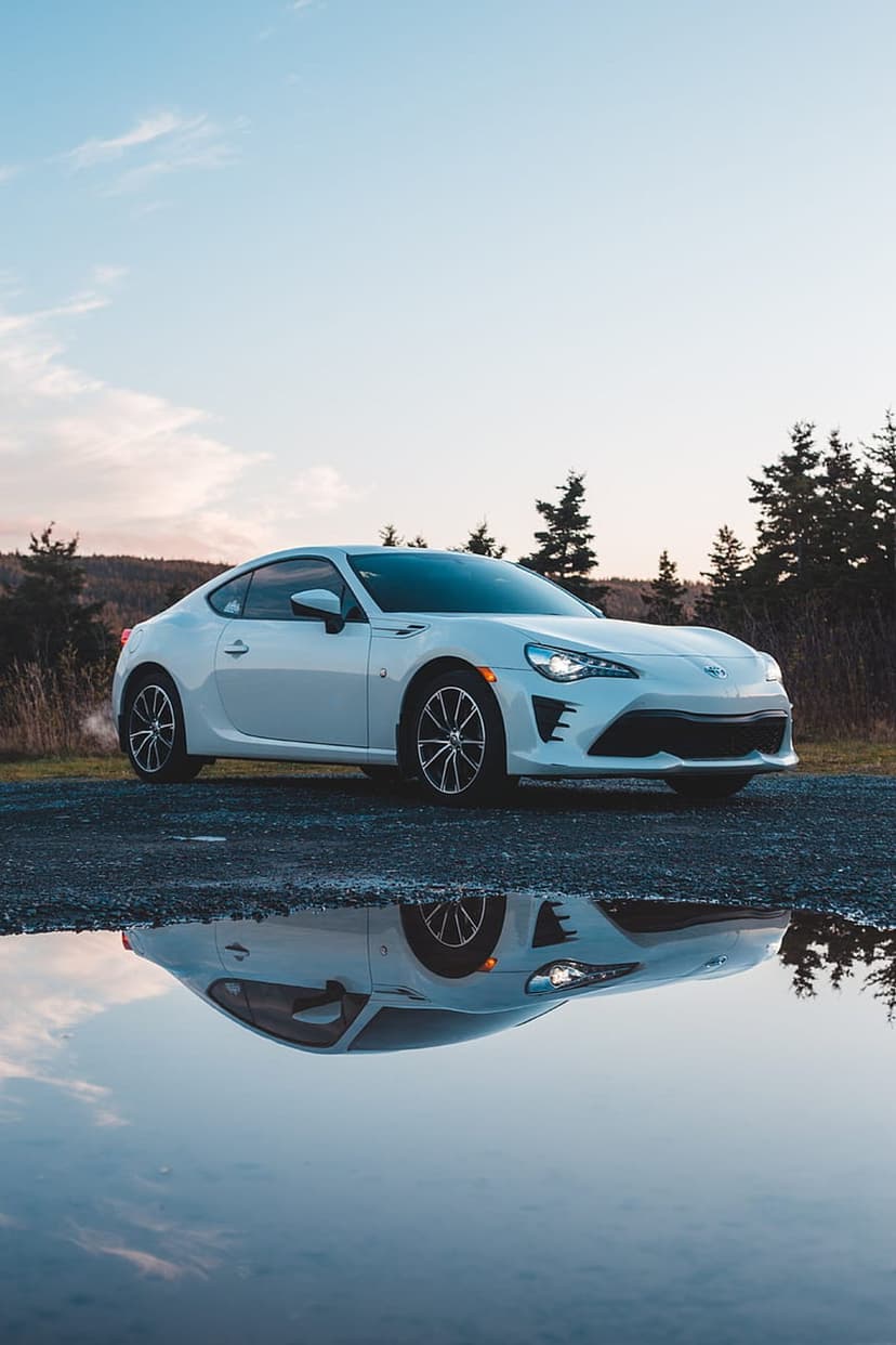 White Toyota 86 sports car reflects in a ground puddle during a colorful sunset amidst towering pine trees.