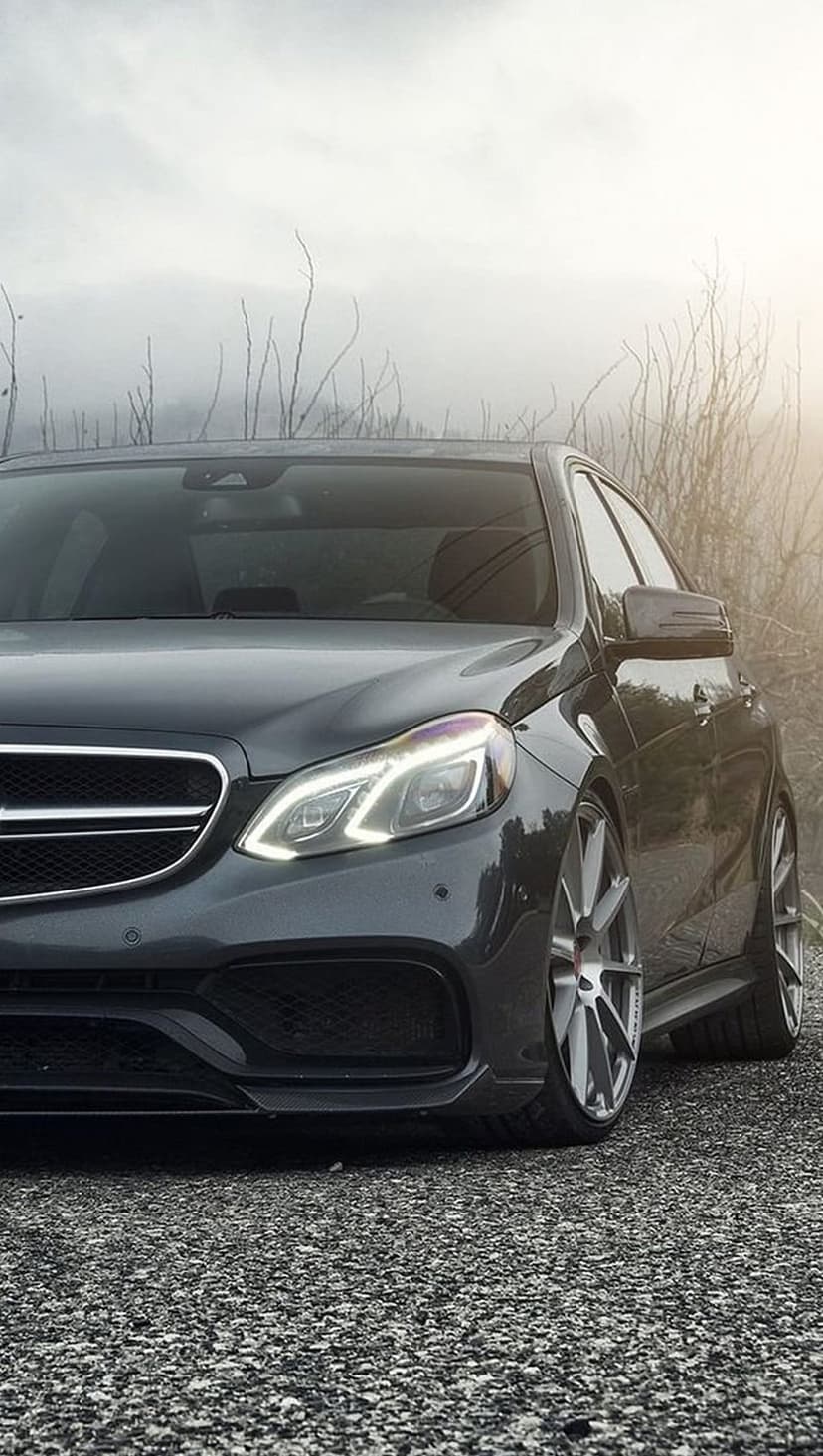 Mercedes E-Class sedan with glowing headlights on a gravel road under a foggy, overcast sky with custom wheels.