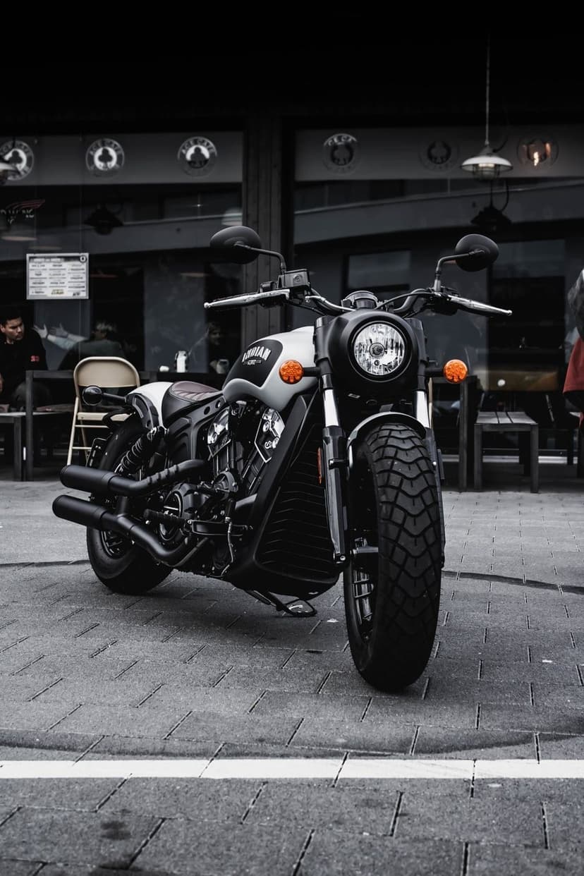 Indian Scout motorcycle with dual exhausts parked on a sidewalk in front of a cafe with large glass windows.
