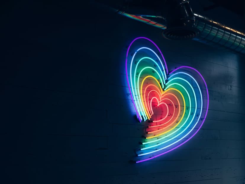 Concentric rainbow neon hearts glow with vibrant multi-colored light against a dark, textured masonry wall.