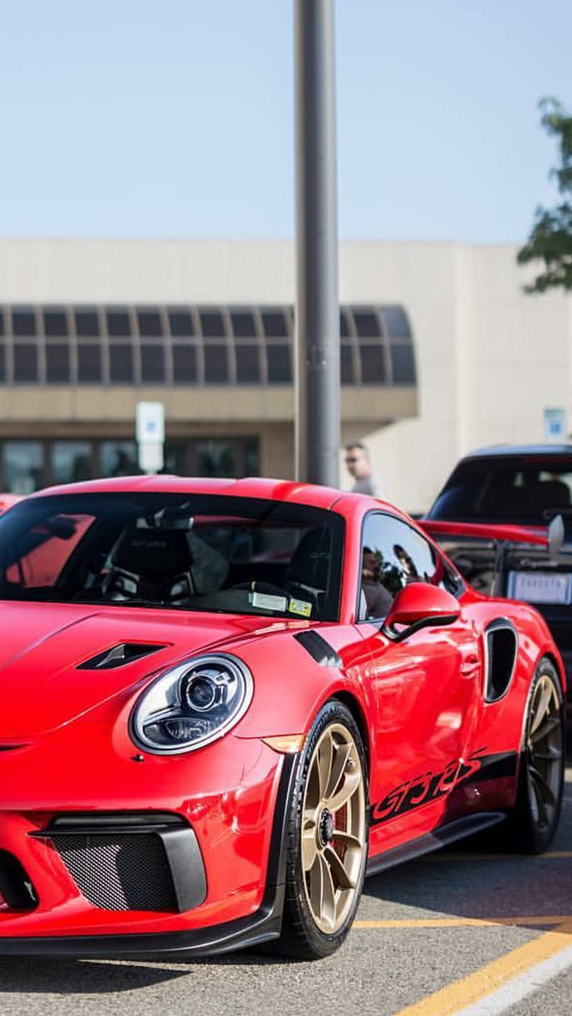 Red Porsche 911 GT3 RS with gold wheels parked on asphalt against a backdrop of trees and a modern building.