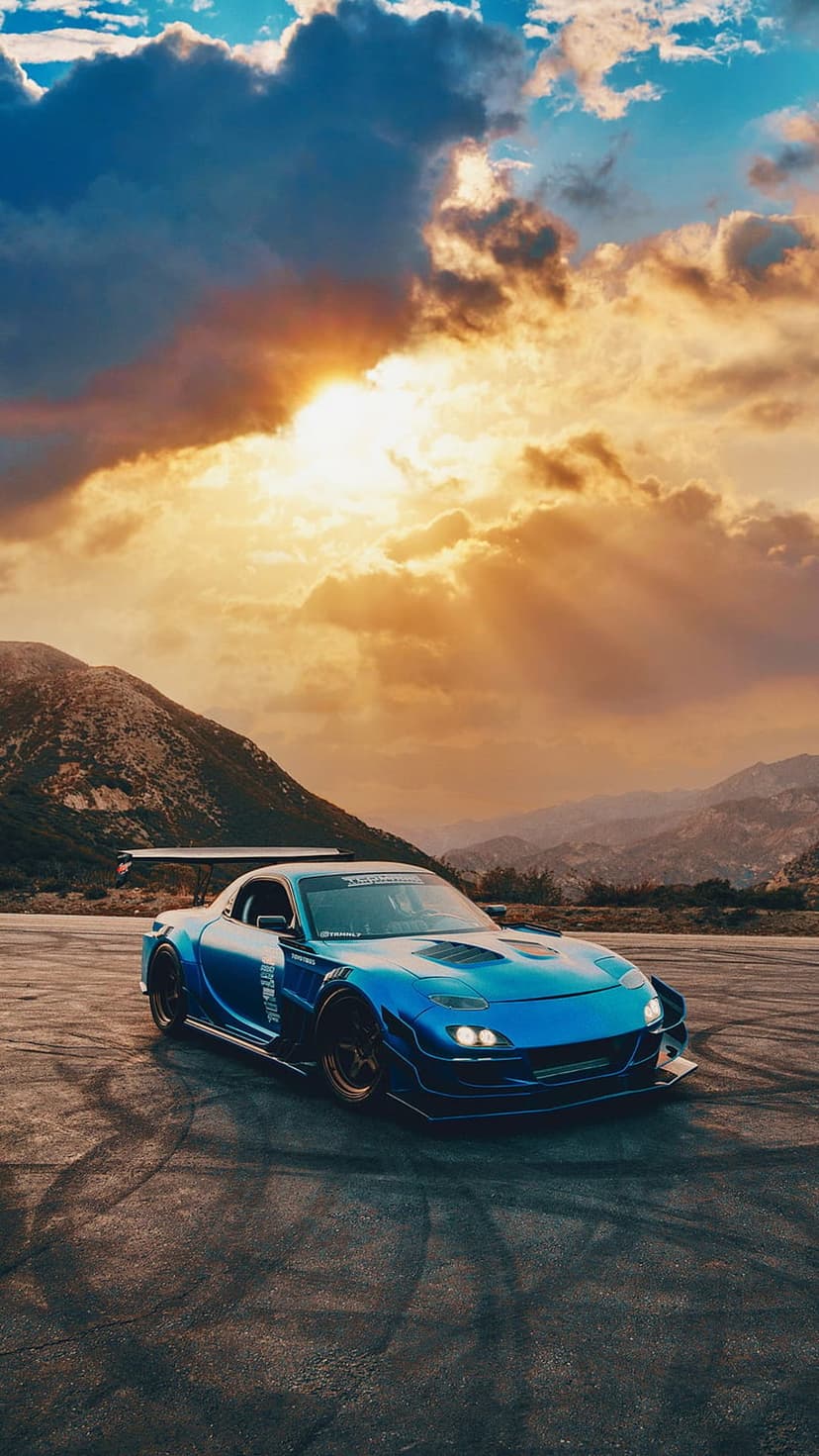 Blue sports car drifting on asphalt with tire smoke under a dramatic orange sunset and mountain range.