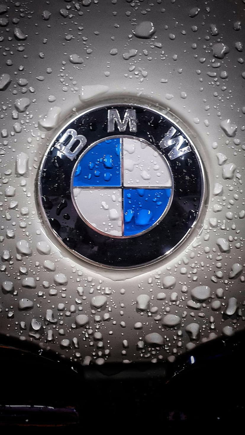 BMW emblem on a silver car hood covered in clear water droplets with soft reflections from an overcast sky.