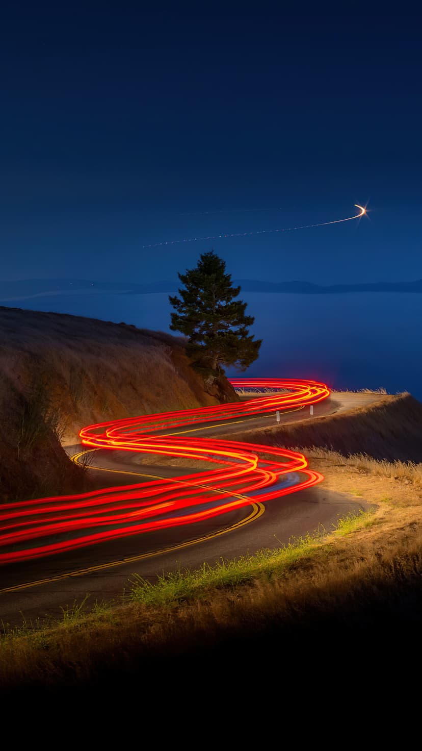 Red vehicle light trails wind along a steep grassy cliff beside a dark ocean under a clear starry night sky.