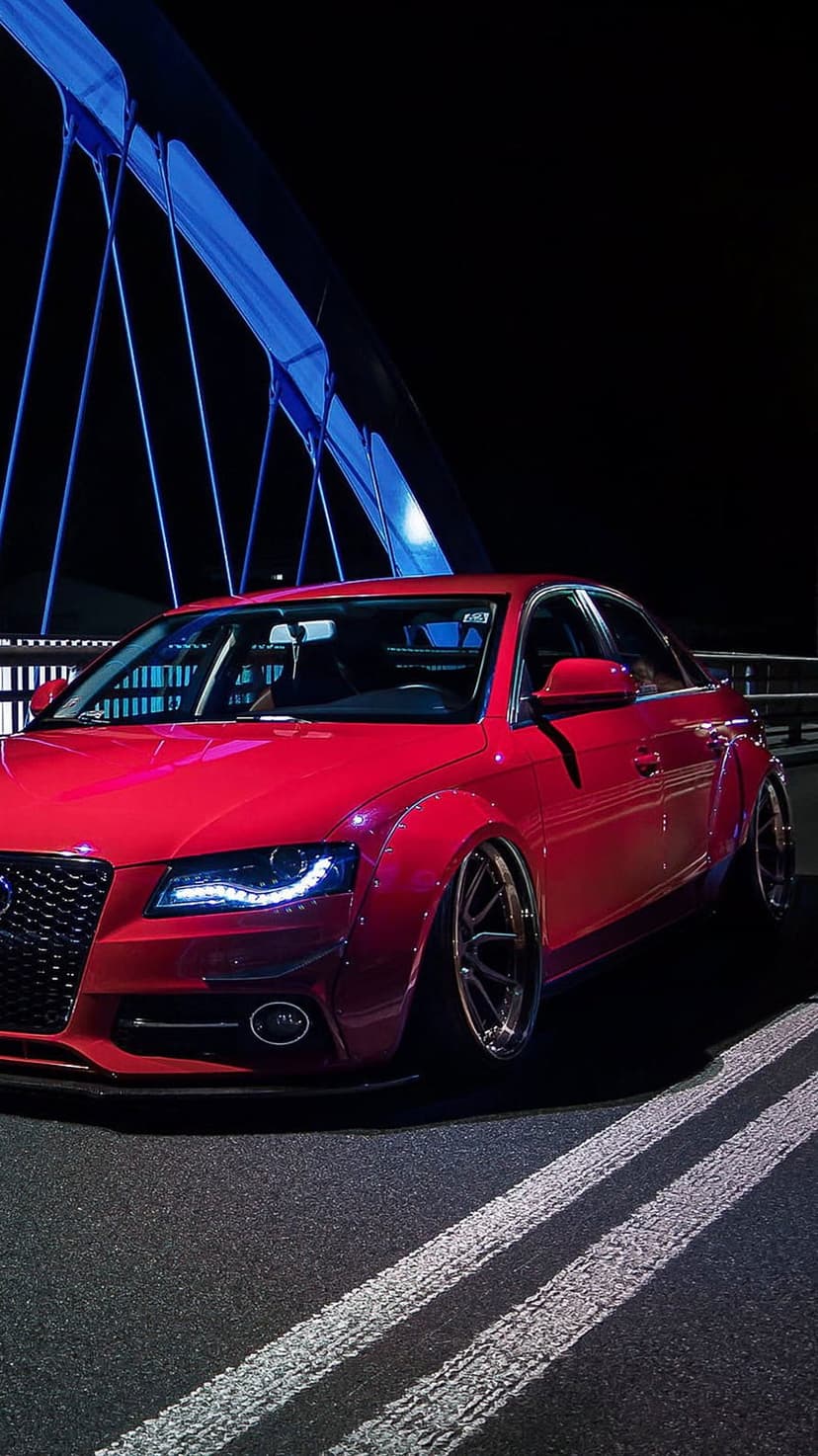 Red Audi A4 sedan with widebody fenders parked on a bridge at night under blue architectural lighting.