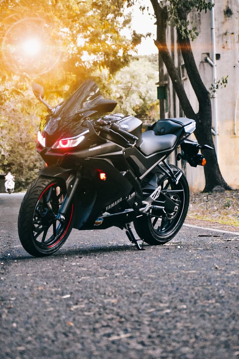Black Yamaha motorcycle with glowing red headlights on an asphalt road surrounded by sunlit trees and greenery.