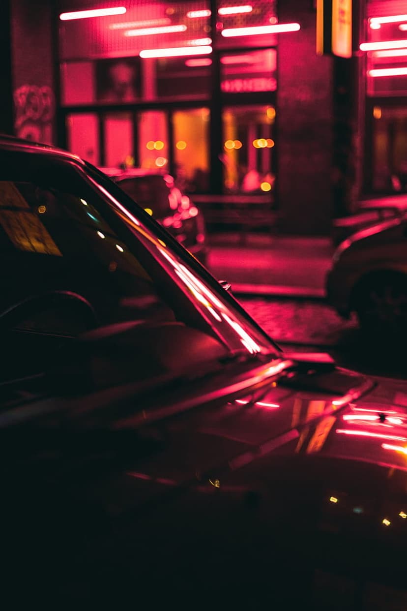 Polished car hood reflecting pink neon lights against a blurred urban backdrop under a dark night sky.