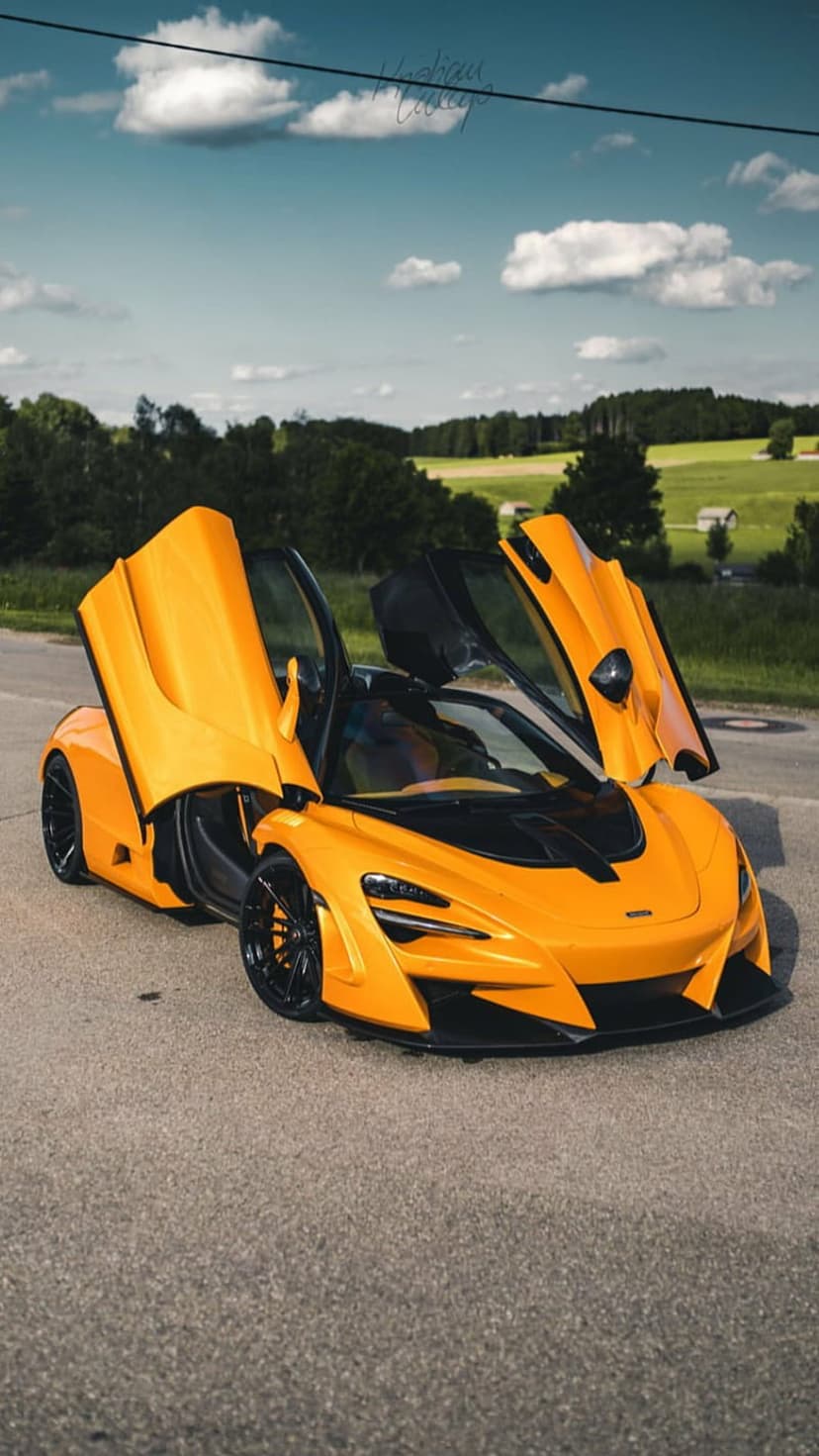 Orange McLaren 720S with dihedral doors open on a rural road against green trees under a cloudy sky.
