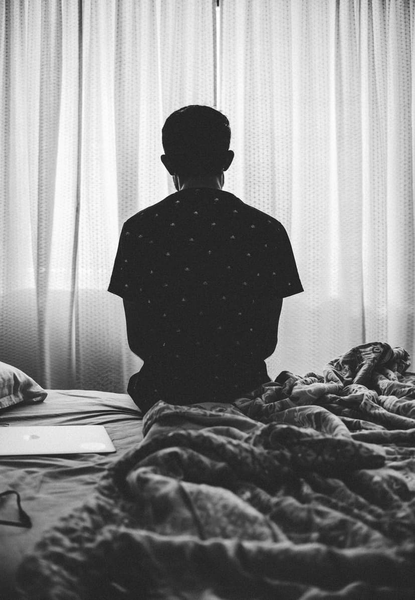 Man sits in silhouette on a rumpled bed facing a bright window with sheer curtains and a nearby laptop.