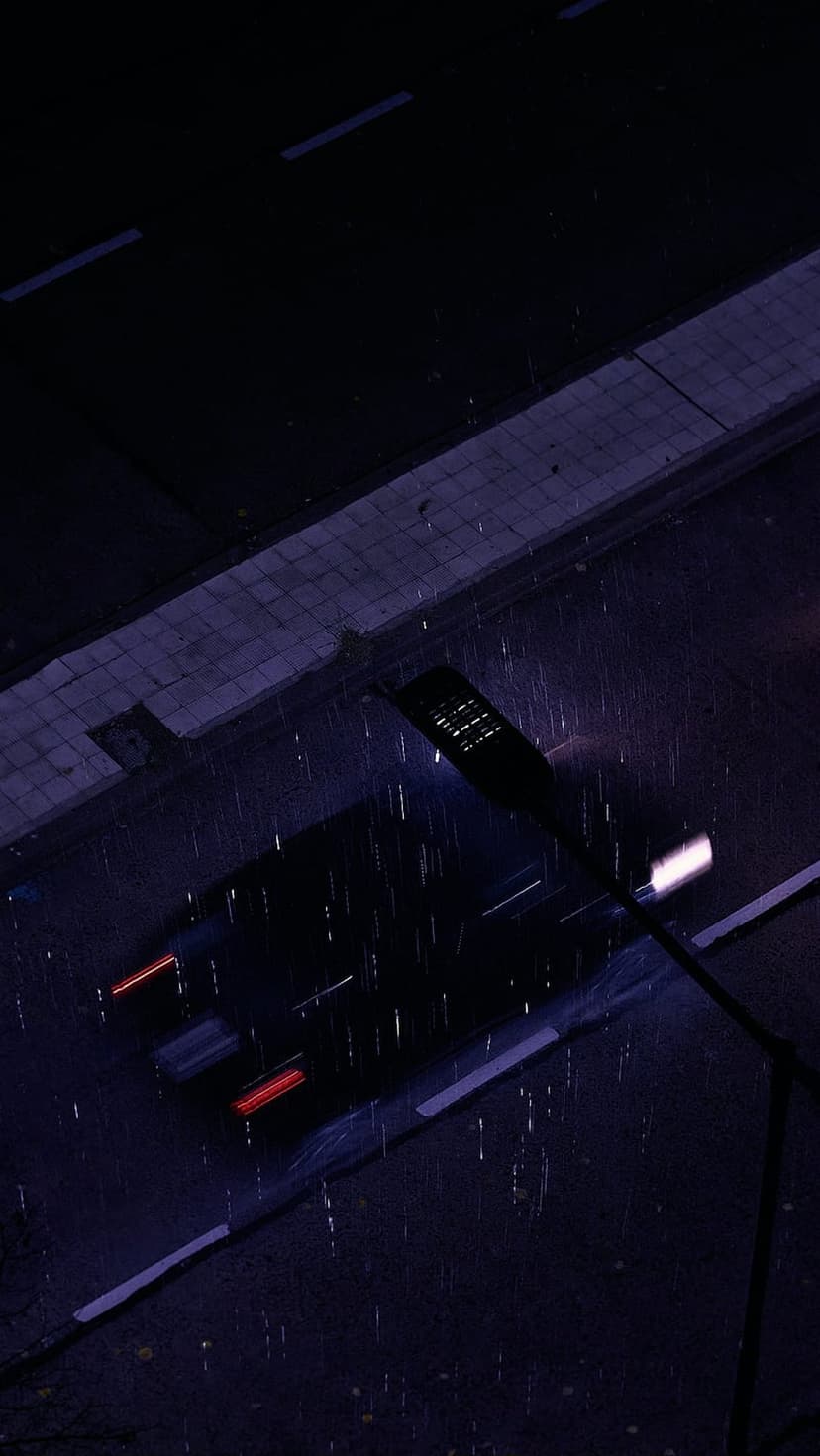 Red and white light trails extend over wet asphalt on a dark rainy street viewed from a high aerial angle.
