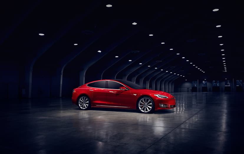 Red luxury electric sedan parked in a dark vaulted warehouse with overhead lights reflecting on a polished floor.