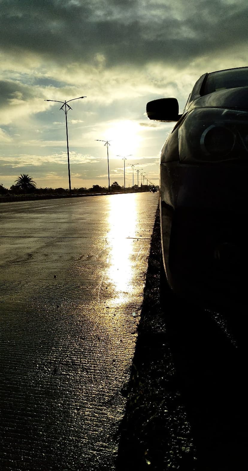 Dark car silhouette parked on a wet, reflective road beneath a dramatic orange and purple sunset with streetlights.