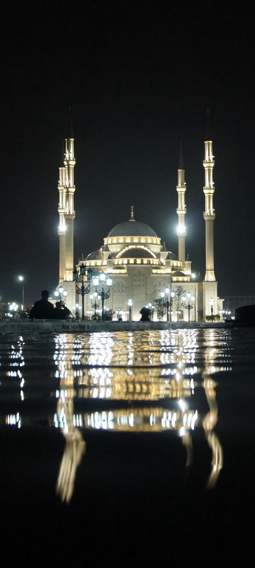 Grand mosque with illuminated golden minarets and dome reflects on a wet surface with silhouetted people nearby.