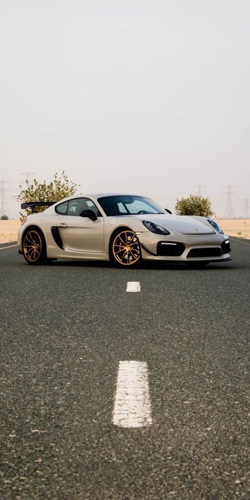 Silver Porsche Cayman GT4 with gold wheels parked on a desert road with power lines under a hazy pale sky.