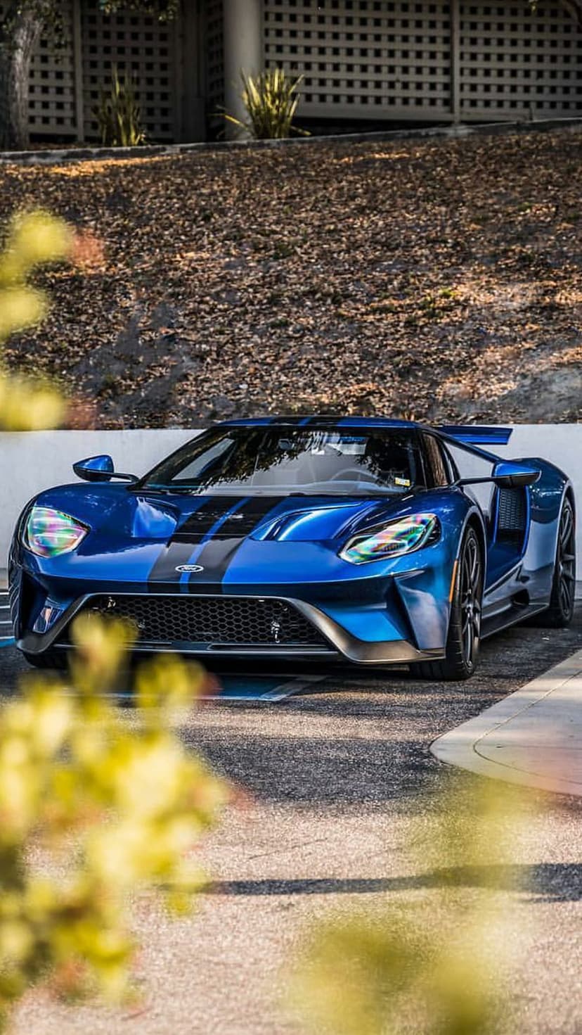 Blue Ford GT supercar with black racing stripes parked outdoors near a building behind blurred yellow leaves.