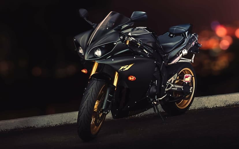 Matte black Yamaha R1 sportbike with gold wheels and forks parked at night with orange bokeh lights.