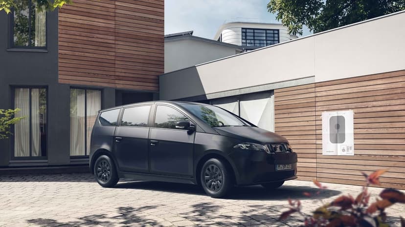 Black Sono Sion solar electric car parked in front of a modern wood-paneled house on a paved driveway in daylight.