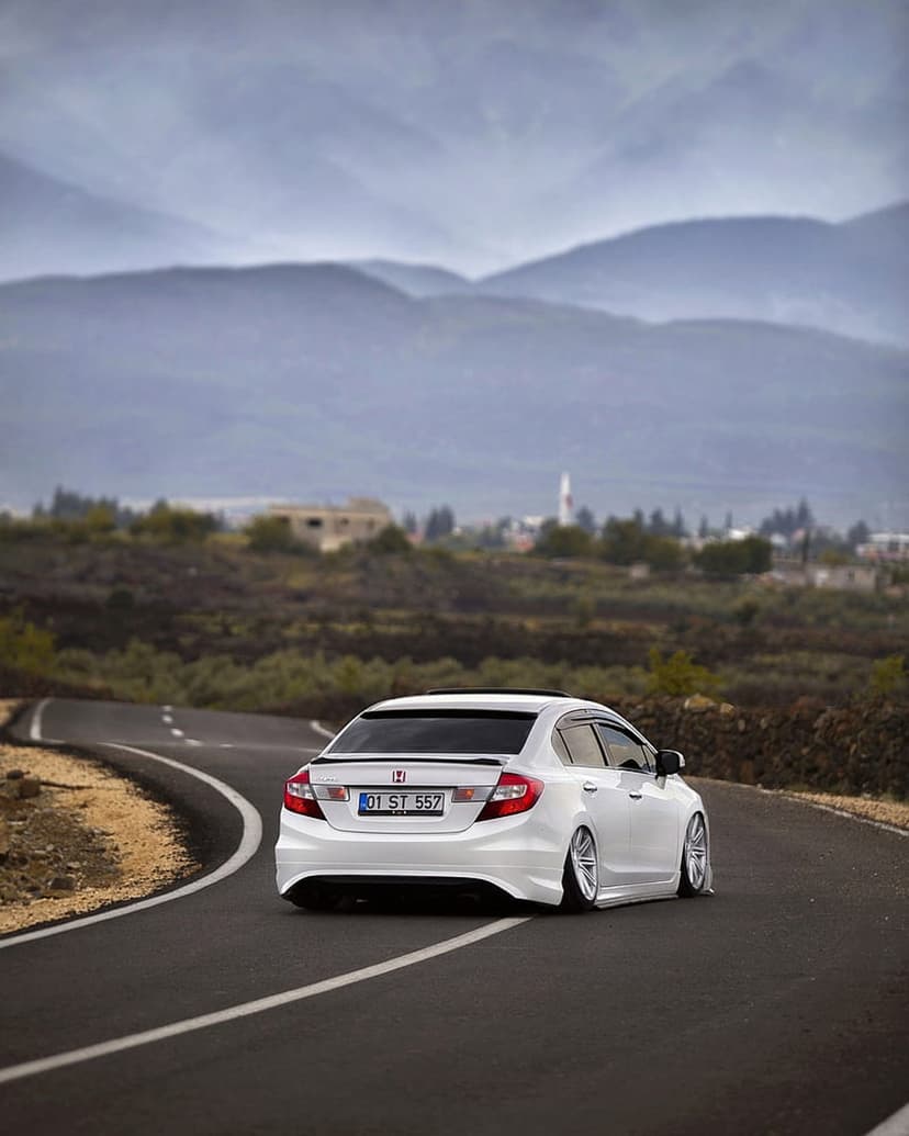 White Honda Civic with custom wheels driving on a winding mountain road with hazy peaks and a valley village.