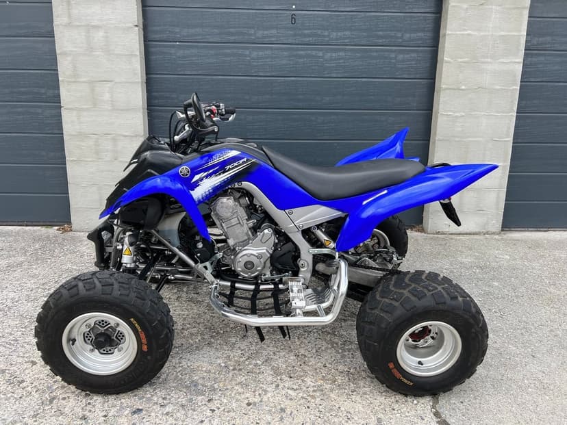 Blue Yamaha Raptor 700R sport ATV parked against a grey garage door with silver engine details and nerf bars.