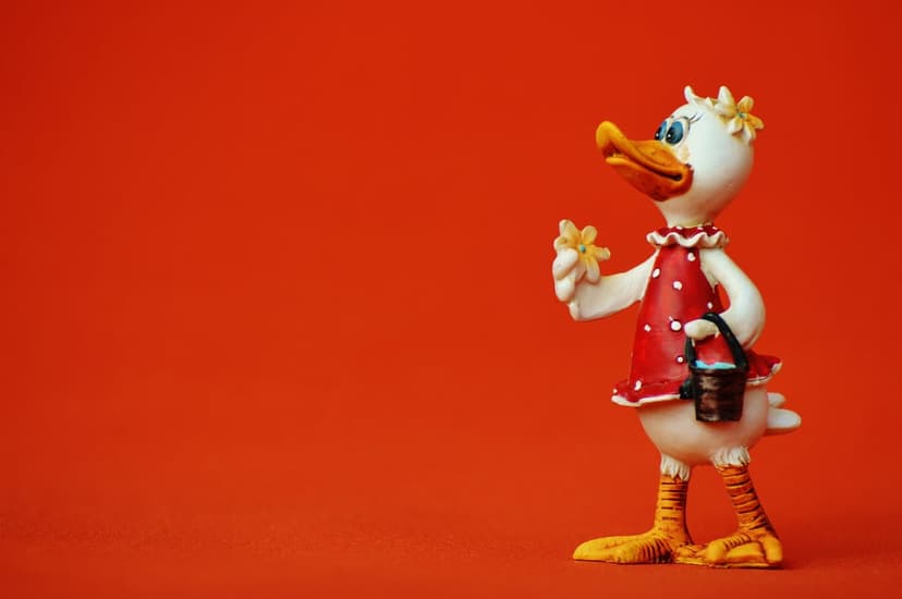 White duck figurine wearing a red polka-dot dress and yellow flowers on a solid bright orange background.