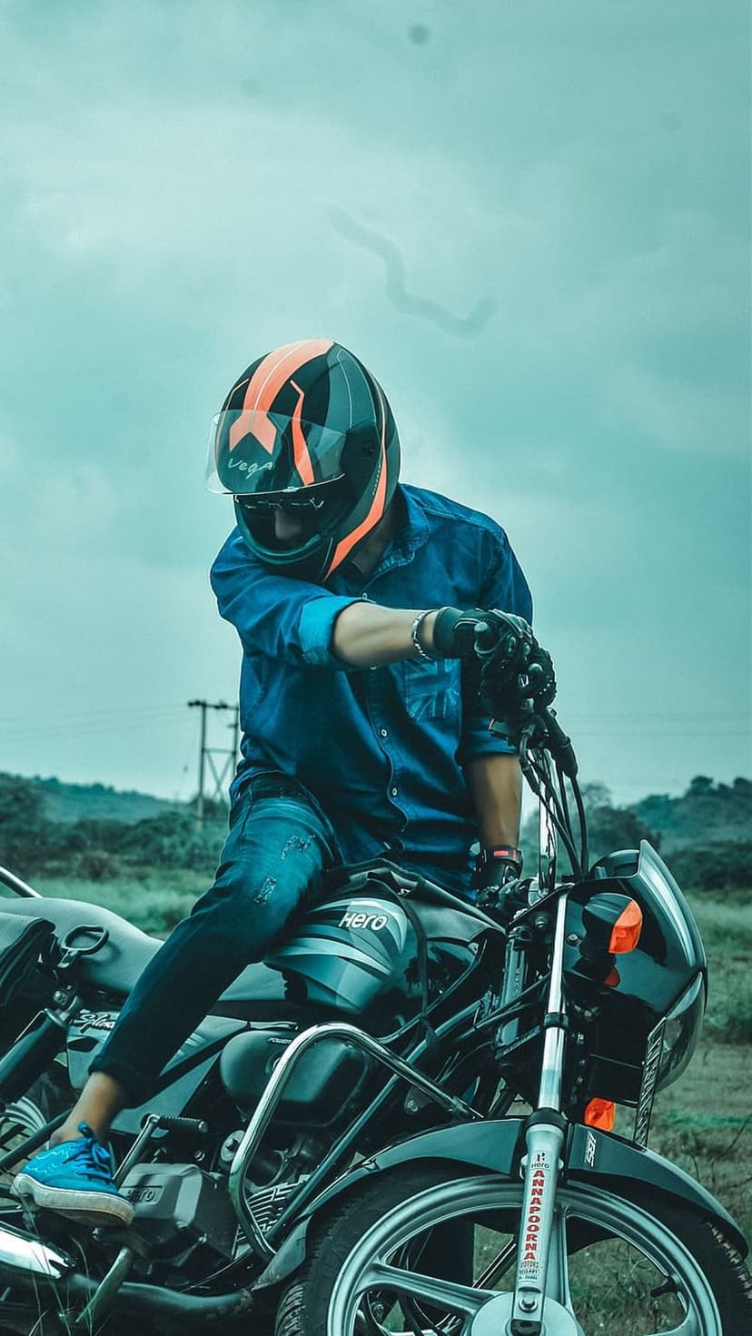 Biker wearing a black and orange helmet sits on a chrome motorcycle against a blurred green landscape background.