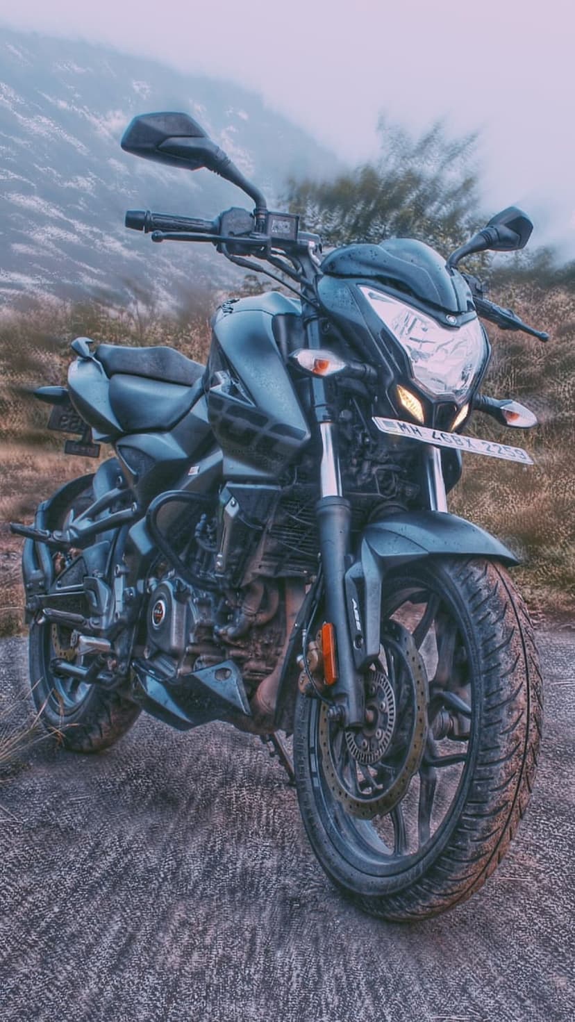 Black motorcycle with glowing lights parked on a gravel mountain road surrounded by thick white mist and tall trees.