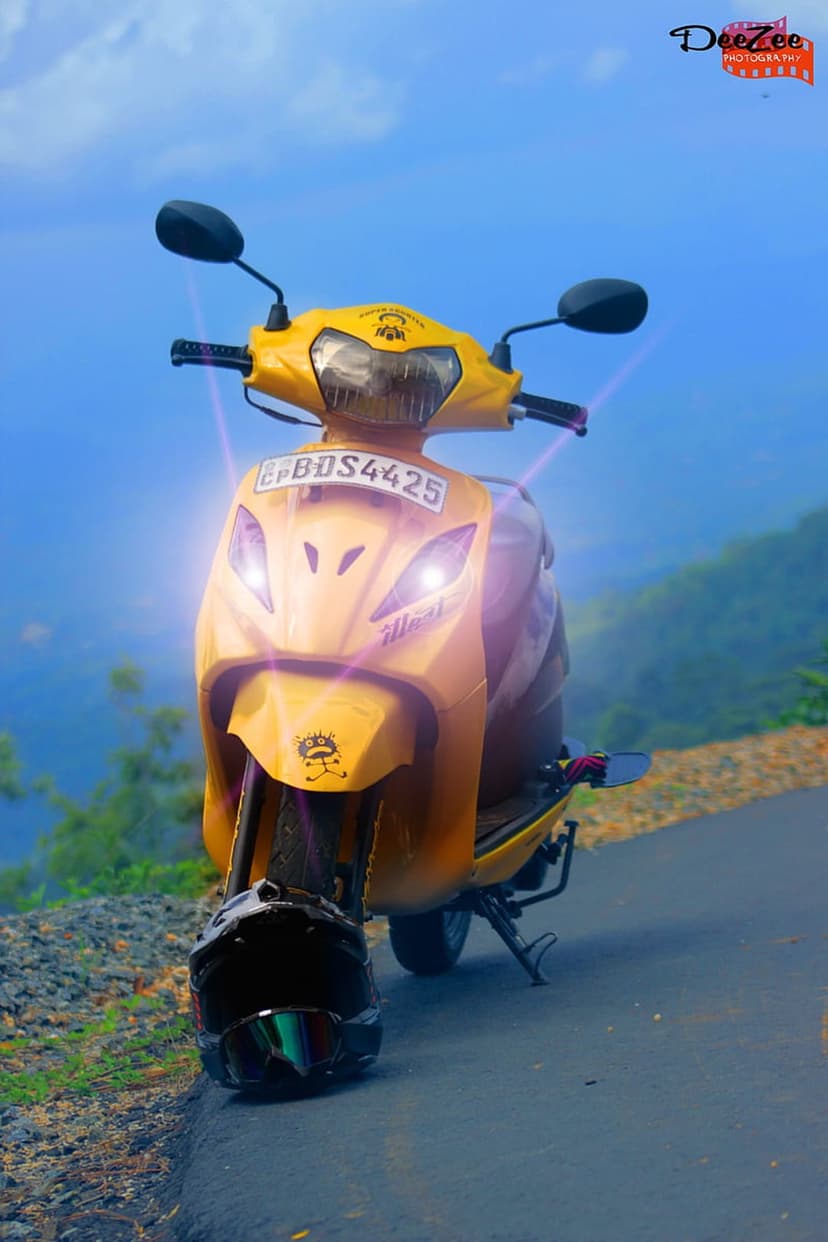 Yellow scooter with glowing headlights parked on a mountain road curve under a vast blue sky with green hills.