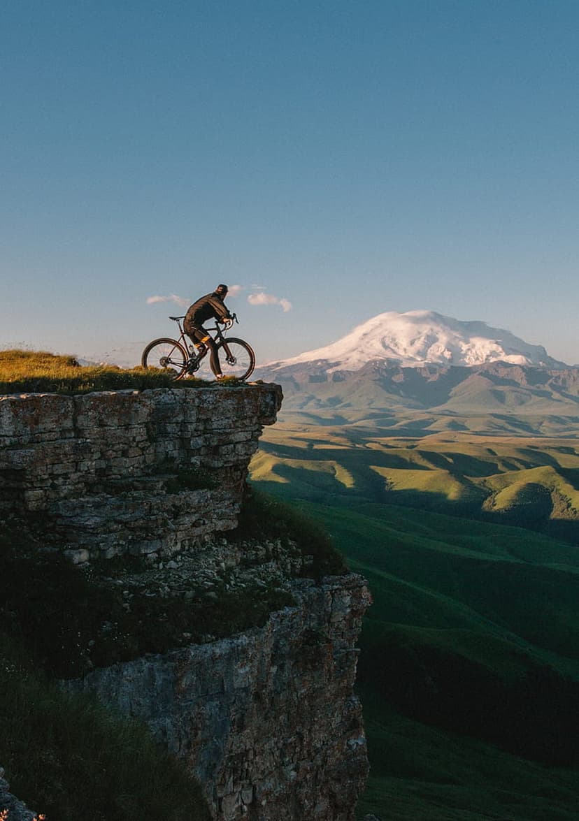 Mountain biker standing on a rocky precipice overlooking a green valley and a massive snow-capped peak under blue sky