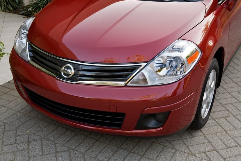 Close-up of a red Nissan car's front grille, chrome logo, and clear headlight on a brick pavement.