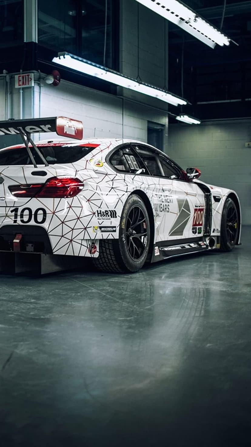 BMW M4 DTM race car with geometric patterns and a large rear wing parked in a brightly lit indoor garage area.