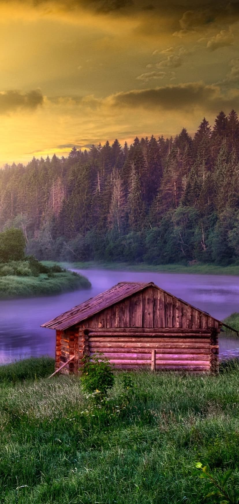 Rustic wooden cabin on a grassy riverbank during a misty orange sunrise with dense evergreen trees in the distance.