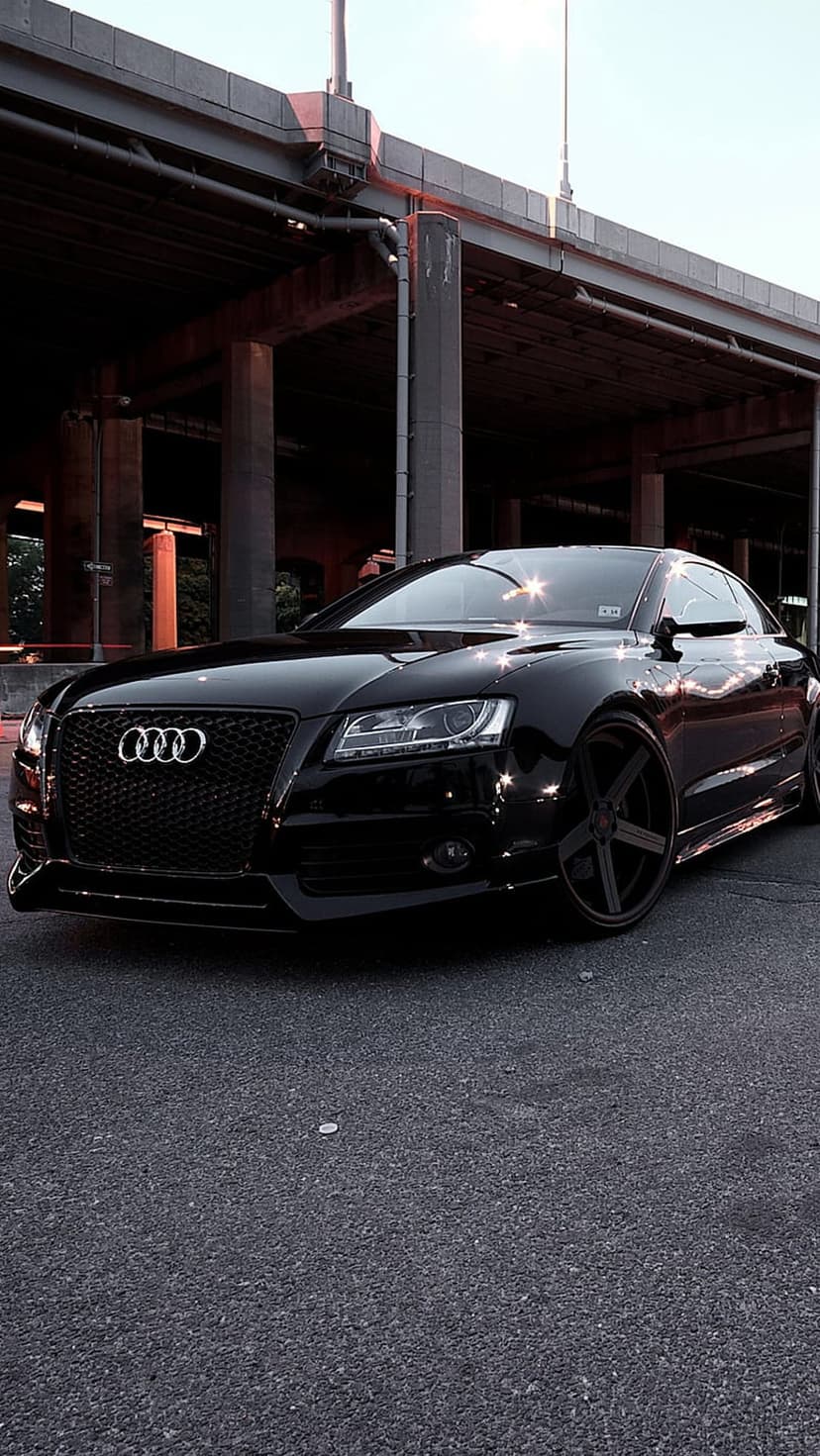 Black Audi coupe with custom rims parked on asphalt under a massive concrete bridge with industrial lighting.