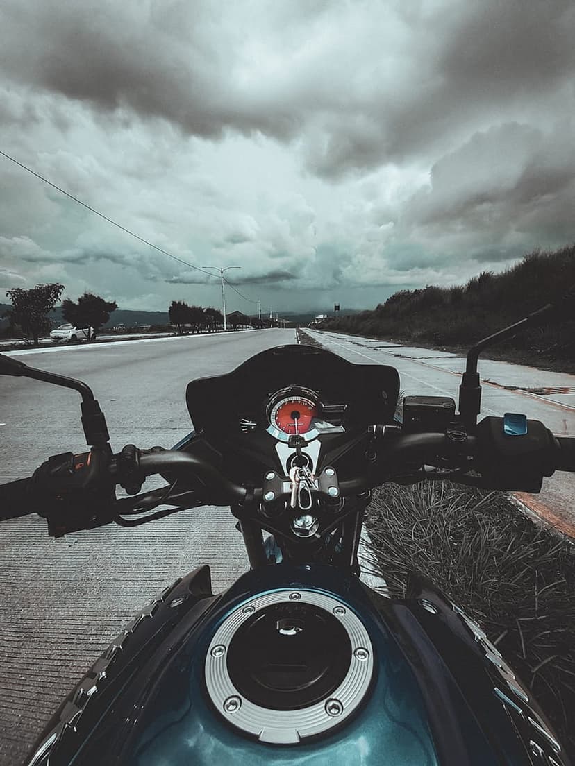Motorcycle handlebars and a red-lit speedometer face a dark, stormy sky on an empty rural road during a journey.