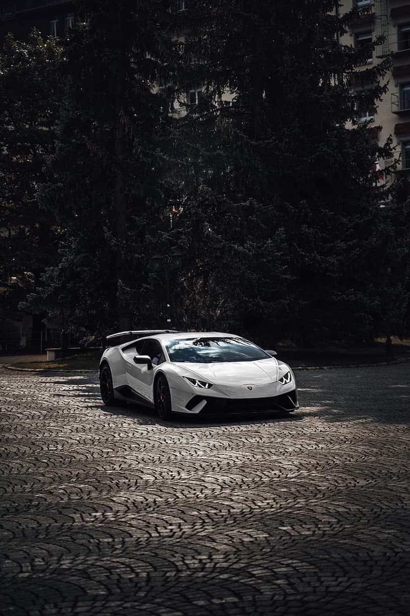 White Lamborghini Huracan supercar parked on textured cobblestones with dark evergreen trees in the distance.