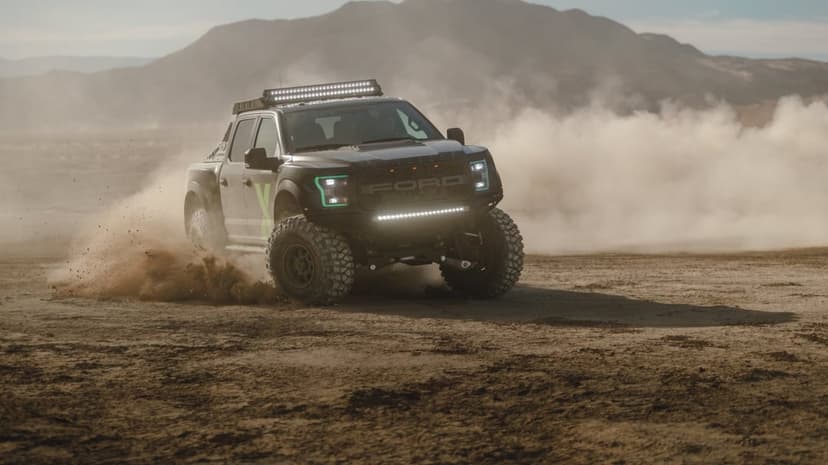 Black Ford Raptor truck drifting on desert sand with bright LED lights and dust clouds against mountains.