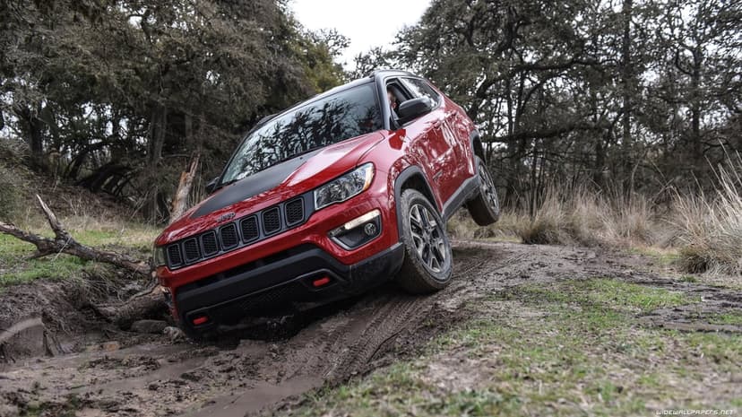 Red Jeep Compass Trailhawk crawling through a muddy rutted forest trail with one wheel lifted in the air.