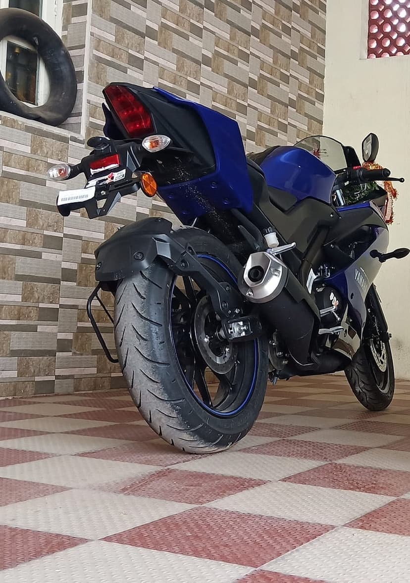 Blue Yamaha R15 motorcycle parked on a checkered floor with its rear tire and exhaust visible against a brick wall.
